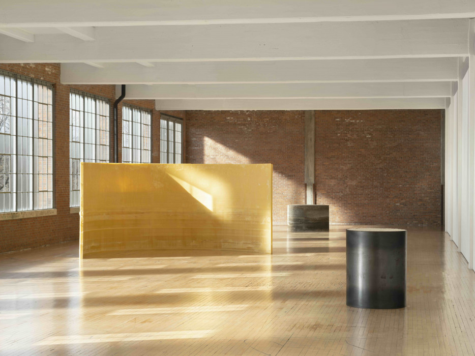 A large curved wall of yellow-tan semi translucent material is in front of three large windows and slight curves towards the camera. To the right of the curved wall, two cylinders made of metal sit in front and behind of the wall. The windows show a blue sunny day, which cast daylight on the floor, curved wall, and metal round